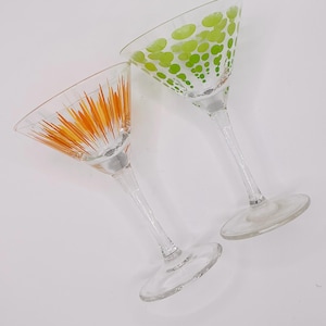 May include: Two clear glass martini glasses with long stems and circular bases. One glass features a vibrant orange sunburst pattern of thin lines, while the other glass displays a pattern of lime green polka dots in varying sizes.