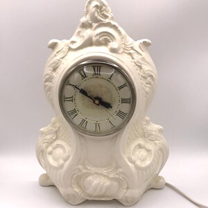 May include: A white ceramic clock with an ornate design. The clock face has Roman numerals and a black hour and minute hand.