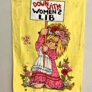 May include: Yellow vintage towel with a cartoon girl holding a sign that reads "DOWN WITH WOMEN'S LIB." The girl wears a pink dress and a floral crown. A butterfly and flowers decorate the towel.