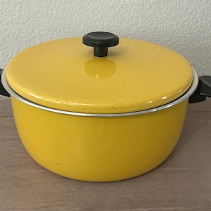 May include: A yellow enamel pot with a matching lid and black handles. The pot has a black knob on the lid and a silver rim. The pot is sitting on a wooden surface.