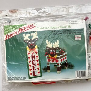 May include: A plastic canvas kit featuring a reindeer centerpiece basket and a matching doorknobber. The kit includes yarn, needles, and a chart. The packaging features the text "Love'n Stitches" and "Christmas plastic canvas kit".