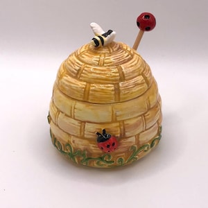 May include: A ceramic honey pot shaped like a beehive with a bee on top and a ladybug on the side. The pot has a red and black dipper with a red ladybug on the end.