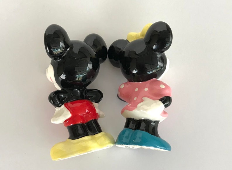May include: A ceramic figurine set of Mickey Mouse and Minnie Mouse. Mickey is wearing red shorts and yellow shoes. Minnie is wearing a pink polka dot dress and blue shoes.