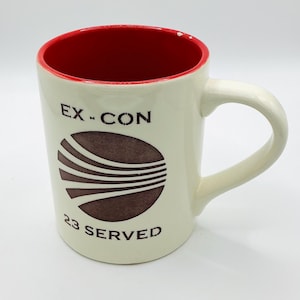 May include: Cream-colored ceramic mug with a red interior. The mug features the text "EX-CON" above a brown, stylized design and the words "23 SERVED" below. The handle is curved and the mug is on a white background.