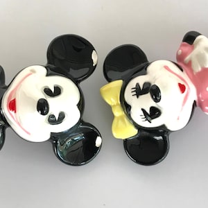 May include: A pair of ceramic Mickey Mouse and Minnie Mouse figurines. Mickey is wearing red shorts and black shoes. Minnie is wearing a pink dress with white polka dots and a yellow bow.