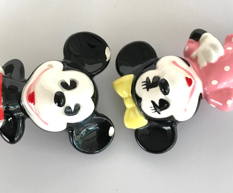 May include: A pair of ceramic Mickey Mouse and Minnie Mouse figurines. Mickey is wearing red shorts and black shoes. Minnie is wearing a pink dress with white polka dots and a yellow bow.