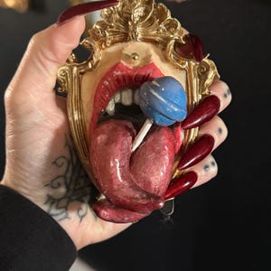 May include: A decorative, ornate gold-framed mirror featuring a stylized open mouth with red lips and tongue, holding a blue lollipop. The mirror has a vintage aesthetic.