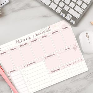 Può includere: Un'agenda settimanale rosa con sezioni per ogni giorno della settimana, preparazione dei pasti e monitoraggio delle abitudini. L'agenda include il testo "Weekly planner" e ha un design morbido e femminile. Sono visibili anche una penna rosa, un mouse per computer e una tastiera.