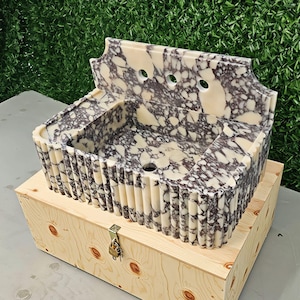 May include: A rectangular marble sink with a cream and dark gray pattern, set on a wooden crate. The sink has a fluted front and a decorative back with three holes. The crate is made of light-colored wood.