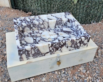 Puede incluir: Un lavabo rectangular de mármol con un diseño blanco y morado oscuro. El lavabo está colocado sobre una caja de madera de color claro con un cierre metálico y un pequeño candado. Es un accesorio de baño.