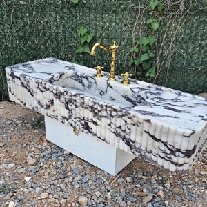 Puede incluir: Un lavabo rectangular de m&aacute;rmol con grifo y accesorios dorados. El lavabo tiene una superficie con vetas blancas y grises y un borde acanalado. Est&aacute; montado sobre un mueble blanco, adecuado para un ba&ntilde;o.