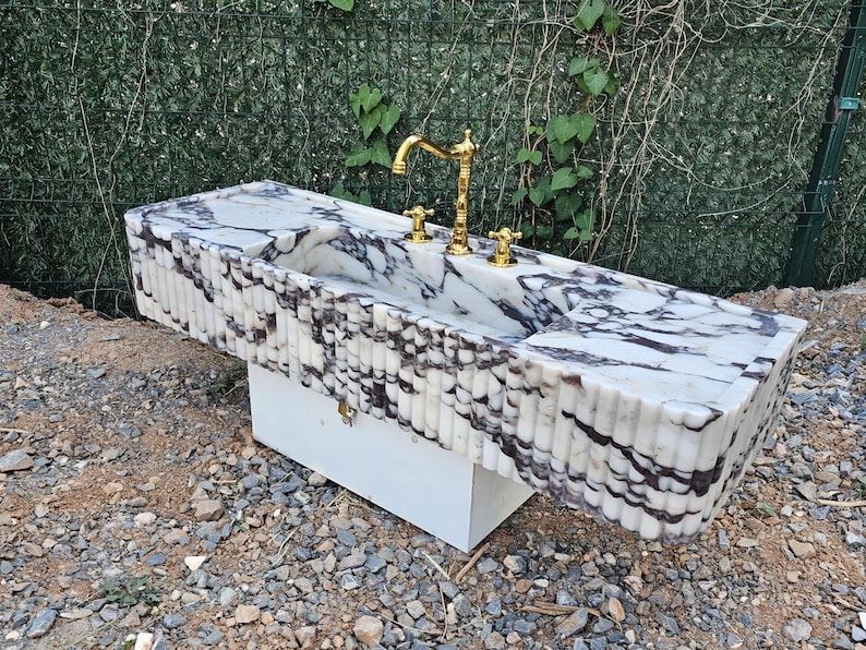 Puede incluir: Un lavabo rectangular de m&aacute;rmol con grifo y accesorios dorados. El lavabo tiene una superficie con vetas blancas y grises y un borde acanalado. Est&aacute; montado sobre un mueble blanco, adecuado para un ba&ntilde;o.