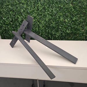 May include: Gray metal shelf brackets with pre-drilled holes, arranged on a white surface against a green backdrop. The brackets are rectangular with a flat, matte finish, designed for wall mounting.