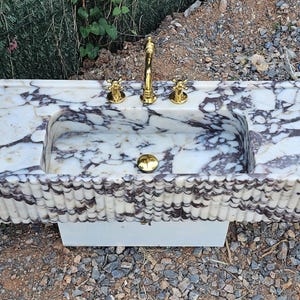 Puede incluir: Un lavabo rectangular de m&aacute;rmol con un patr&oacute;n veteado blanco y gris oscuro. El lavabo tiene una pila integrada, grifo y accesorios dorados. La parte delantera del lavabo tiene un dise&ntilde;o estriado. El lavabo est&aacute; sobre una base blanca.
