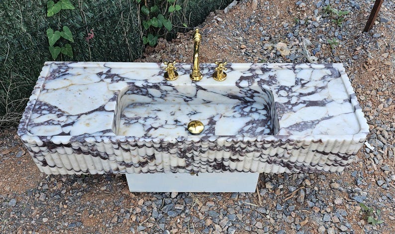 Puede incluir: Un lavabo rectangular de m&aacute;rmol con un patr&oacute;n veteado blanco y gris oscuro. El lavabo tiene una pila integrada, grifo y accesorios dorados. La parte delantera del lavabo tiene un dise&ntilde;o estriado. El lavabo est&aacute; sobre una base blanca.