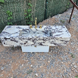 Puede incluir: Un lavabo rectangular de m&aacute;rmol con un patr&oacute;n veteado blanco y morado. El lavabo tiene un grifo dorado y dos manijas doradas. La base del lavabo es blanca y el lavabo est&aacute; al aire libre.