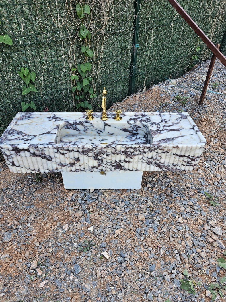 Puede incluir: Un lavabo rectangular de m&aacute;rmol con un patr&oacute;n veteado blanco y morado. El lavabo tiene un grifo dorado y dos manijas doradas. La base del lavabo es blanca y el lavabo est&aacute; al aire libre.