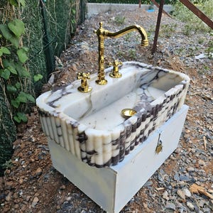 Könnte beinhalten: Ein rechteckiges Waschbecken aus Marmor mit weiß-dunkelvioletter Maserung und geriffeltem Außendesign. Das Waschbecken ist mit einer polierten goldfarbenen Armatur, zwei passenden goldfarbenen Griffen und einem goldfarbenen Ablauf ausgestattet.