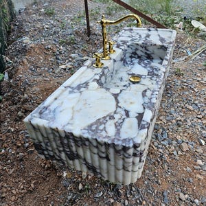 Puede incluir: Un lavabo rectangular de m&aacute;rmol con una superficie estampada en blanco y gris. El lavabo tiene un grifo y accesorios dorados. Los lados del lavabo tienen un dise&ntilde;o acanalado.
