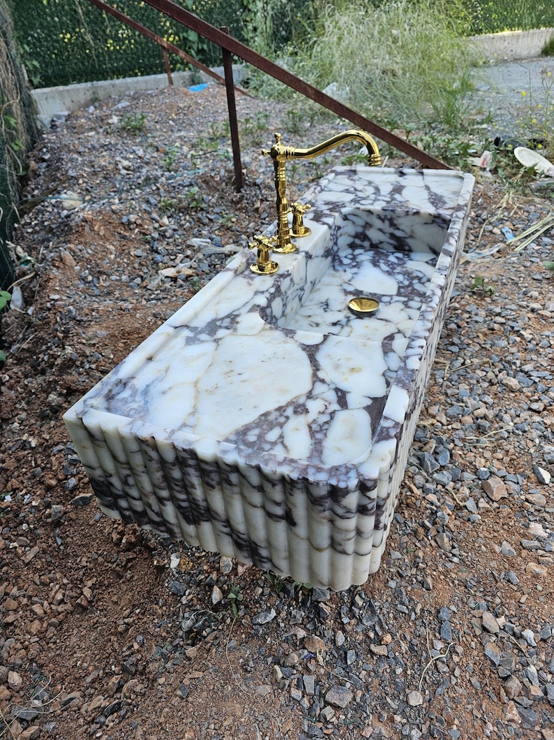 Puede incluir: Un lavabo rectangular de m&aacute;rmol con una superficie estampada en blanco y gris. El lavabo tiene un grifo y accesorios dorados. Los lados del lavabo tienen un dise&ntilde;o acanalado.
