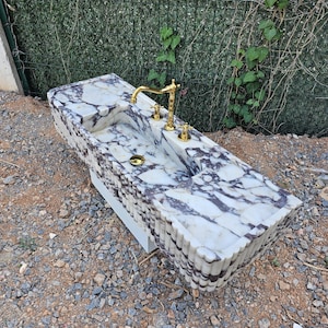 Puede incluir: Un lavabo rectangular de m&aacute;rmol con un patr&oacute;n veteado blanco y morado. El lavabo tiene un grifo y accesorios dorados. El lavabo est&aacute; sobre una base blanca y est&aacute; al aire libre.