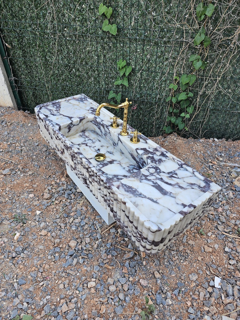 Puede incluir: Un lavabo rectangular de m&aacute;rmol con un patr&oacute;n veteado blanco y morado. El lavabo tiene un grifo y accesorios dorados. El lavabo est&aacute; sobre una base blanca y est&aacute; al aire libre.