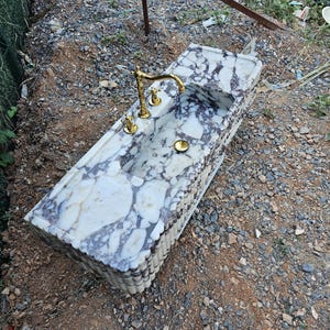 Puede incluir: Un lavabo rectangular de m&aacute;rmol con grifo y accesorios dorados. El lavabo tiene una superficie con un patr&oacute;n blanco y morado. El grifo est&aacute; centrado sobre la pila, con dos manijas a cada lado. El lavabo est&aacute; al aire libre.