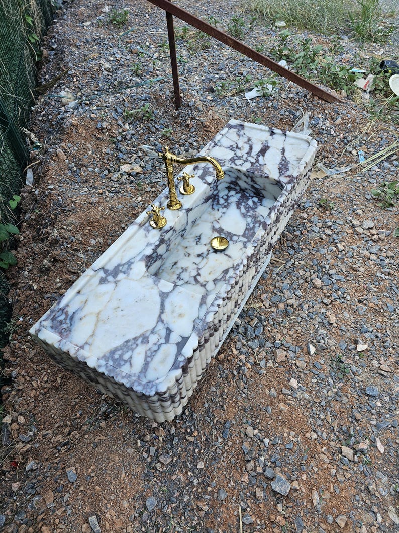Puede incluir: Un lavabo rectangular de m&aacute;rmol con grifo y accesorios dorados. El lavabo tiene una superficie con un patr&oacute;n blanco y morado. El grifo est&aacute; centrado sobre la pila, con dos manijas a cada lado. El lavabo est&aacute; al aire libre.