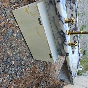 Puede incluir: Un lavabo de m&aacute;rmol con grifos y accesorios dorados. El lavabo tiene un patr&oacute;n veteado blanco y gris. Un mueble de madera de color claro est&aacute; unido al lavabo.