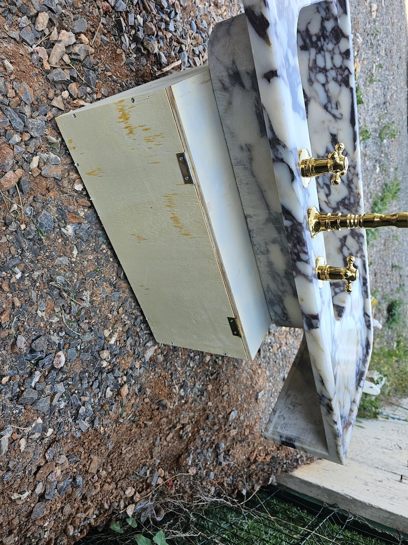 Puede incluir: Un lavabo de m&aacute;rmol con grifos y accesorios dorados. El lavabo tiene un patr&oacute;n veteado blanco y gris. Un mueble de madera de color claro est&aacute; unido al lavabo.