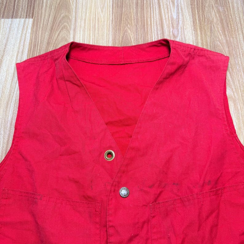 May include: A sleeveless red vest with a V-neck design. The vest has two front pockets and a metal button closure. A metal grommet is visible near the top of the vest. The vest is made of a lightweight fabric.
