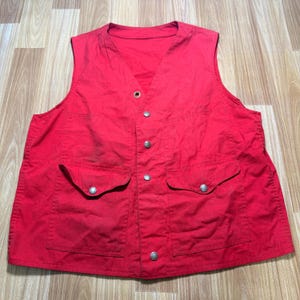 May include: A red vest with a V-neck, two front pockets, and silver-coloured buttons. The vest is sleeveless and appears to be made of a durable fabric. The vest has a classic design.