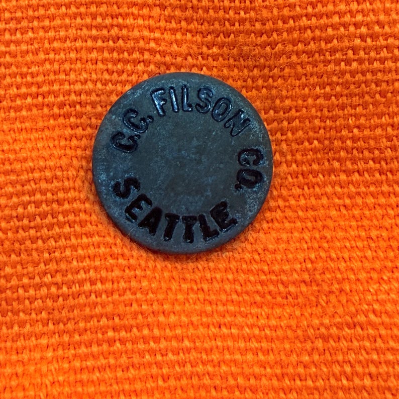 May include: A dark gray, circular button with the text "C.C. FILSON CO. SEATTLE" embossed around the edge. The button is set against a textured orange fabric background.