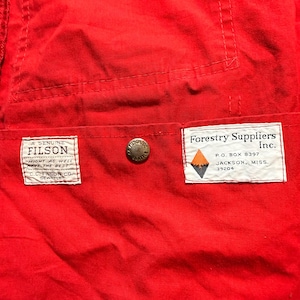 May include: Close-up of a vibrant red fabric featuring two rectangular labels and a metal button. One label reads "A Genuine Filson", the other says "Forestry Suppliers Inc." with an address.