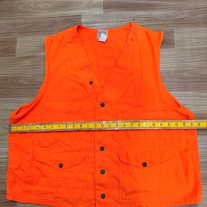 May include: Bright orange hunting vest with four front pockets and black button closures. The vest has a V-neck design and is sleeveless. A measuring tape is placed across the vest, indicating a width of approximately 26 inches.