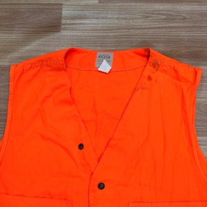 May include: Bright orange vest with two black buttons and two pockets. The vest has a V-neck design and a small white tag with the brand name "FILSON". The vest is made of a durable material and is likely designed for outdoor activities.
