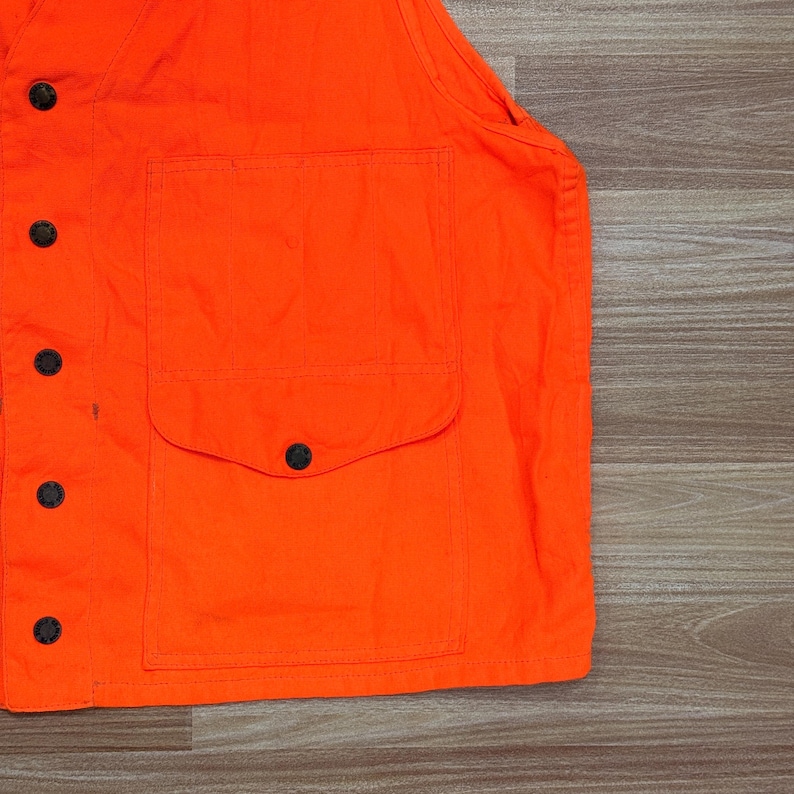May include: Bright orange vest with a large pocket and black buttons. The vest is made of a durable fabric and has a classic design. The vest is likely used for hunting or safety purposes.