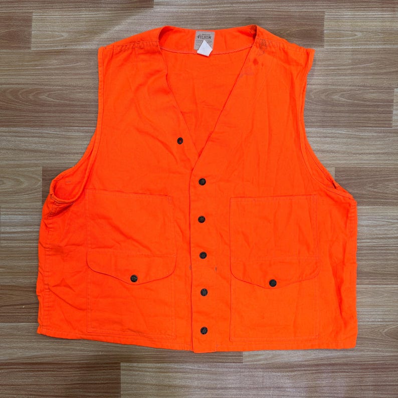 May include: Bright orange vest with a V-neck, two front pockets, and black button closures. The vest has a label at the top and is made of a durable fabric. The vest is designed for outdoor activities.