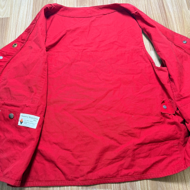 May include: A vibrant red vest featuring several pockets and button fastenings. The vest includes a collar and a label that reads "Forestry Suppliers Inc."