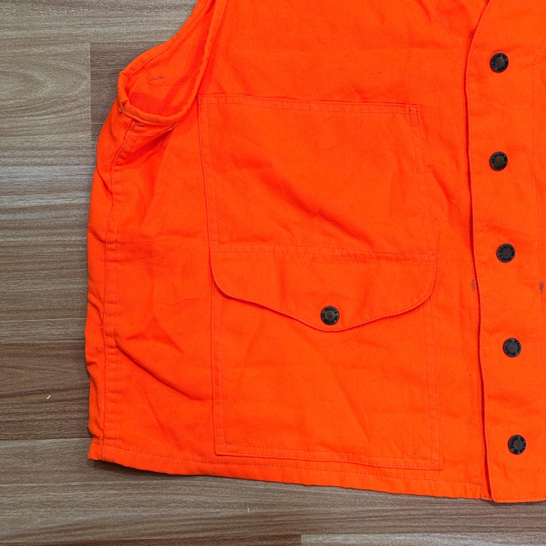 May include: Bright orange vest with a large pocket and black buttons. The vest is made of a durable fabric and has a classic design. A practical garment for outdoor activities.