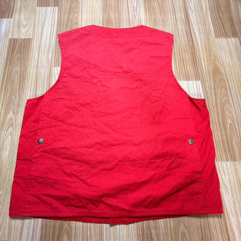 May include: A vibrant red vest with a V-neck. It features two pockets, each secured with a silver button. The material looks lightweight. The vest is displayed flat on a wooden surface.
