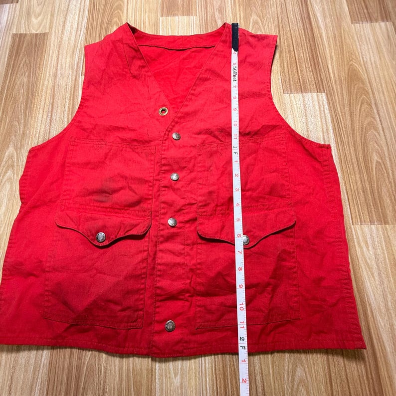 May include: A bright red vest with a V-neck, two front pockets, and silver-colored buttons. The vest is approximately 61 cm long, measured with a tape measure. The vest is made of a sturdy fabric.