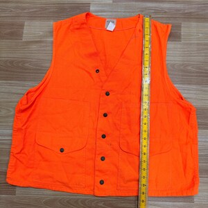May include: Bright orange hunting vest with a V-neck, two front pockets, and black button closures. A yellow ruler is placed next to the vest. The vest is approximately 26 inches long.