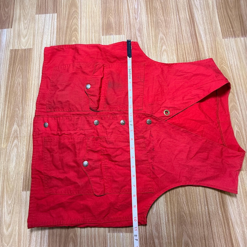 May include: A vibrant red vest featuring several pockets and silver press studs. The vest has a tailored design with a collar and is approximately 61 cm long. The vest is made of a lightweight fabric.