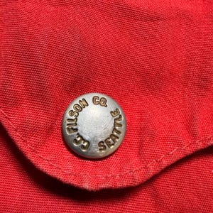 May include: Close-up of red fabric with a silver button. The button is embossed with the words "FILSON CO. SEATTLE" in a circular design. The fabric appears to be a sturdy material, possibly canvas or a similar durable textile.