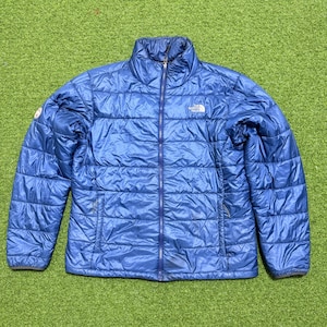 May include: A blue quilted jacket with a full-length zip and a high collar. The jacket features the North Face logo on the chest and a small patch on the left sleeve. The jacket is made of a shiny, lightweight material.