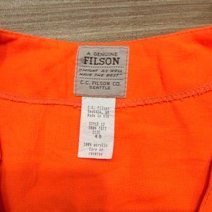 May include: Close-up of a bright orange garment with a Filson label. The label reads "A Genuine Filson" and "Might as well have the best." A smaller tag lists the style, size 48, and material as 100% acrylic. The garment is made by C.C. Filson Co. Seattle.