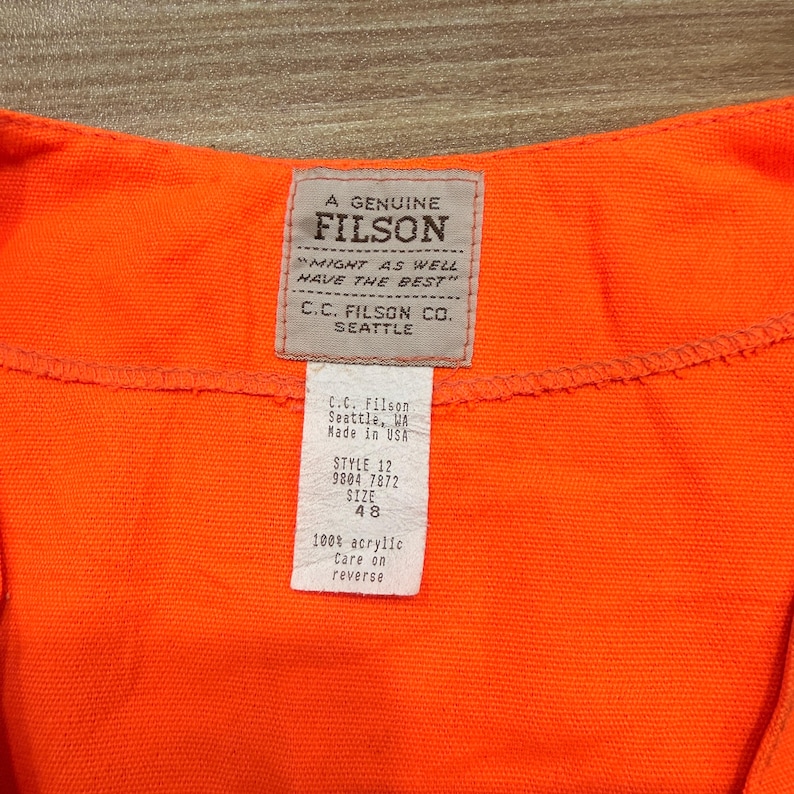 May include: Close-up of a bright orange garment with a Filson label. The label reads "A Genuine Filson" and "Might as well have the best." A smaller tag lists the style, size 48, and material as 100% acrylic. The garment is made by C.C. Filson Co. Seattle.