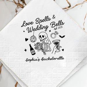 May include: White paper napkin with a black graphic design featuring a skeleton, disco ball, ring, and cocktail glass. Text reads "Love Spells & Wedding Bells est. 2025" and "Sophia's Bachelorette". The napkin is on a white plate with gold accents.