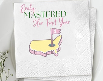 Op de afbeelding: Witte papieren servetten met een ruitpatroon. De servetten hebben de tekst "Emily MASTERED Her First Year" in roze en groen. Er staat ook een gele en roze kaart van de Verenigde Staten met een golfflag op de servet.